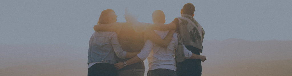 Four people with their arms around each other, standing outside at sunset.