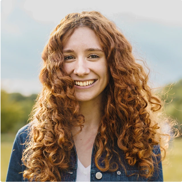 A woman with curly hair standing outside and smiling at the camera.