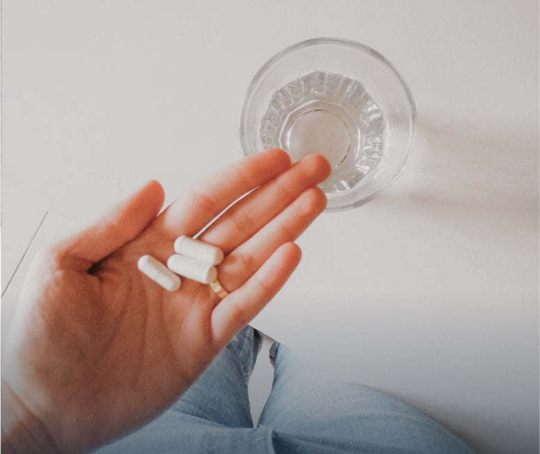 A hand holding three pills, with a glass of water on the table below.