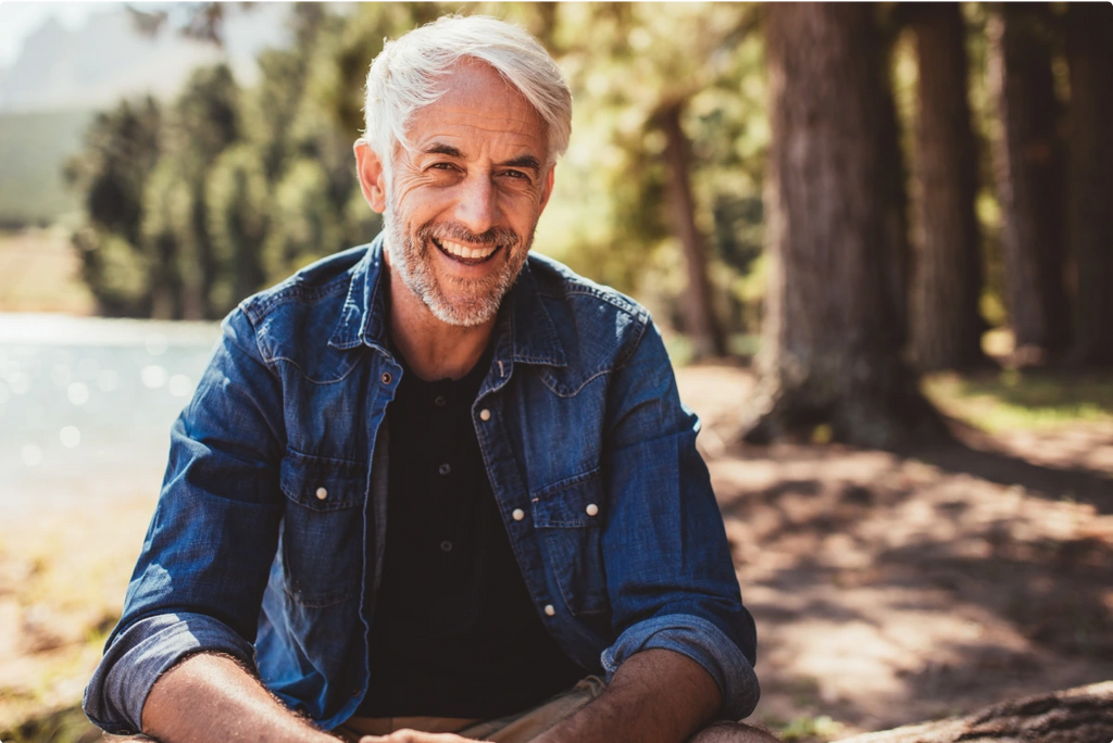 A man with white hair and a denim jacket sitting outside in a forest.