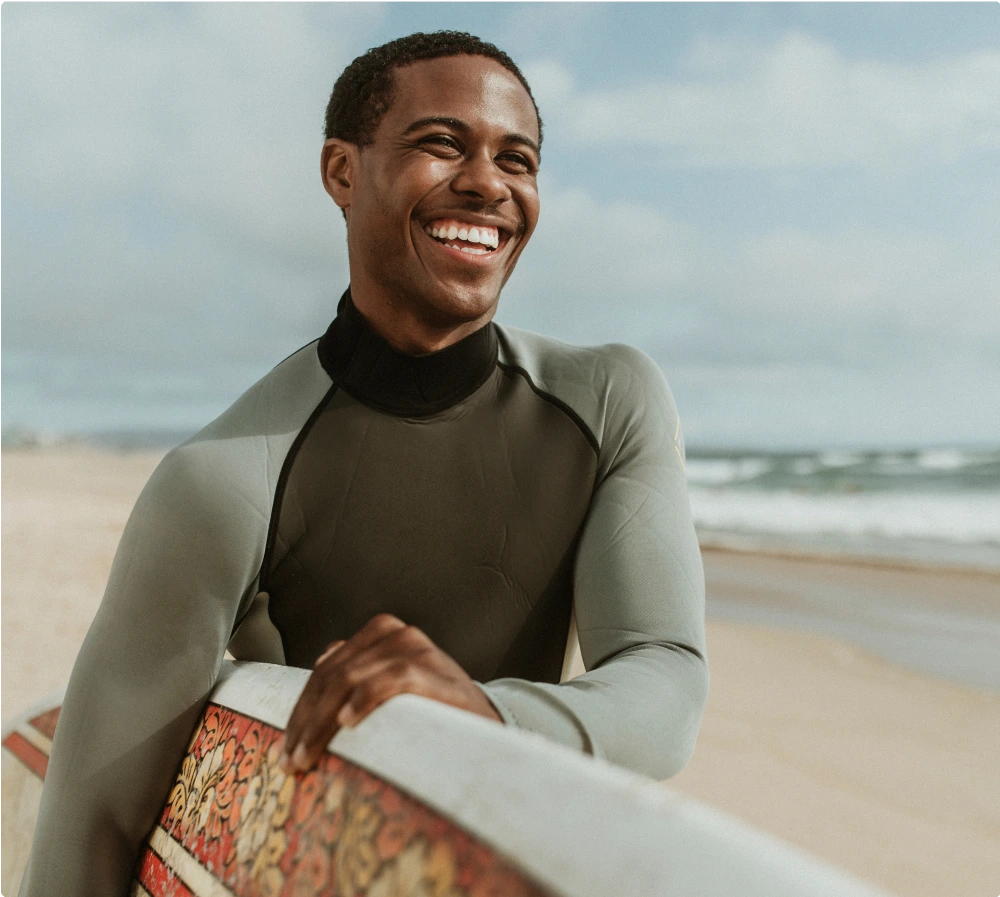 A man in a wetsuit on a beach, holding a surfboard. 